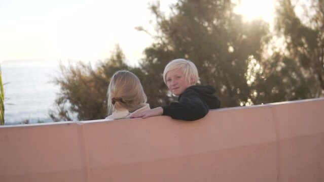 Blond boy sitting on a concrete bench with his younger sister and looking at the sea in the park. Spain