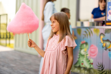 Little happy girl holds pink cotton candy while visiting amusement park during a summer vacation