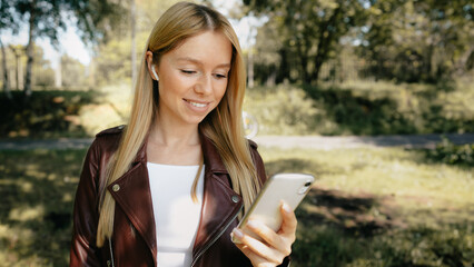 Young caucasian girl with wireless headphones in the park using tablet, phone and smiling