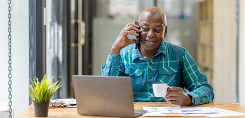 Portrait of Senior African man sitting on laptop at the home office and having a phone conversation with the insurance company. - obrazy, fototapety, plakaty