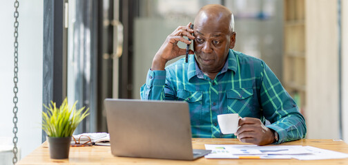Portrait of Senior African man sitting on laptop at the home office and having a phone conversation with the insurance company.