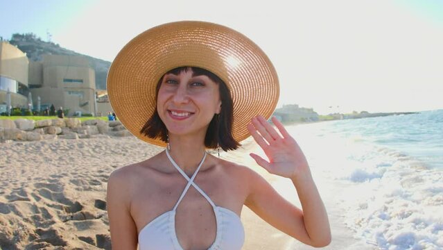 Portrait of a beautiful young woman in a straw hat on the beach. Cheerful young woman smiles and waves at the camera. Happy girl enjoying a trip.