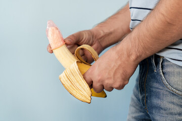 Man's hands are holding banana with elastic condom on it blue background. - obrazy, fototapety, plakaty