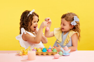 Obraz na płótnie Canvas Beautiful little girls with hair accessories looking at painted and decorated Easter egg against yellow background. Concept of Easter holiday, celebration, traditions, childhood, happiness