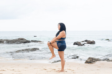 Beautiful, curly-haired, young woman in gym clothes doing stretches on the beach sand.