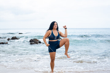 Fitness woman, standing, doing leg exercises facing the camera on the beach sand against the sea and cloudy sky.