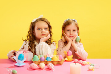 Little cute girls sitting at table with panted, decorated, Easter eggs against yellow background. Concept of Easter holiday, celebration, traditions, childhood, happiness