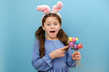 Portrait of charming cheerful preteen girl child in easter bunny ears pointing on small colored eggs, expression enjoy celebration of Christian holiday, posing isolated over blue color background wall
