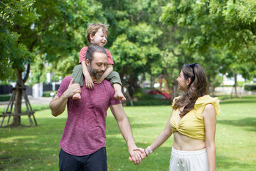 A man and woman are walking together holding hands in a beautiful green park, while the father holds a giggling small child on his shoulder.