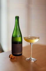 An elegant glass on a long stem with champagne and an open bottle of sparkling wine with a cork. A cup with a cool alcoholic drink on a wooden table in the living room in the house.
