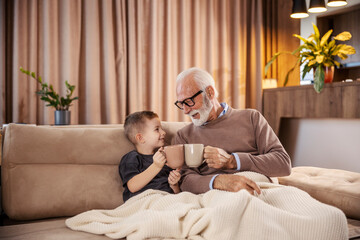 A grandfather and his grandson sitting on sofa at cozy home and toasting with mugs.