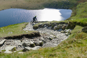 Downhill mountain biking, rider descending steep and rough terrain above glacier lake, Snowdonia,...