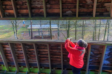Senior female tourist taking photos with her mobile phone at observation tower in Hoge Kempen national park, disused railway track and bare trees in background, sunny day in As, Limburg Belgium