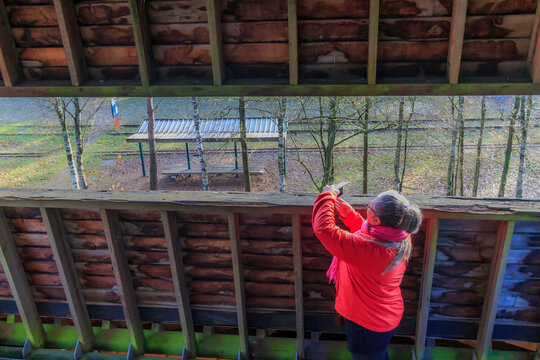 Senior female tourist taking photos with her mobile phone at observation tower in Hoge Kempen national park, disused railway track and bare trees in background, sunny day in As, Limburg Belgium