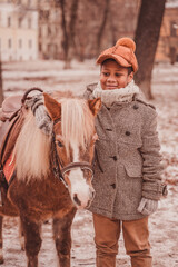 the boy stands next to the pony and hugs the pony by the neck, smiling and looking to the left