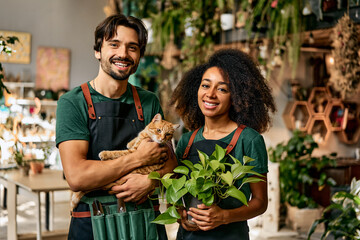 Welcoming touch at shop. Portrait of multiracial family in green uniform posing together and smiling at their own floral boutique. Charming woman holding flower pot while man caressing red fluffy cat.