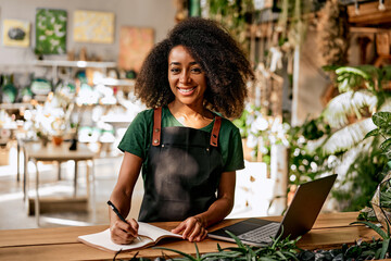 Confident successful sincere smiling charming business woman florist gardener entrepreneur in black apron and green t-shirt in plant shop writing in notebook working on laptop. - obrazy, fototapety, plakaty