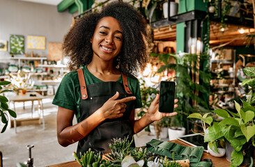 Beautiful smiling successful business lady florist holding phone and pointing finger at it while standing behind shop counter with plants and flowers. Online sales, application, order, delivery.