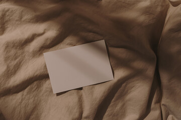 Blank paper sheet card with mockup copy space on crumpled beige bed blanket with soft warm sunlight shadows. Flat lay, top view aesthetic minimal brand template