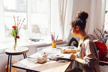Young creative pretty african american girl artist paints with pastels while sitting at table in front of window.