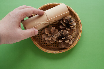 A hand is putting a gift wrap into a wicker basket with pine cones, green background, close-up. Gifts for a holiday, surprise for a holiday