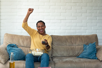 Young African American woman sitting at home on sofa watching sport game on TV, eating popcorn and...