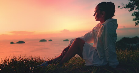 Woman in bright pink sunset light relax on green grass meadow enjoying aerial sea view. Girl in white dress meditate sitting on ground, outdoor lifestyle travel on summer holiday vacation.