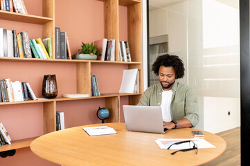 African-American male student in the library, guy using laptop for remote work sitting in the...
