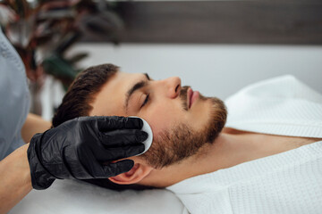 Facial skin cleansing in beauty salon. Beautician in black gloves wipes the face of young man with sponge.
