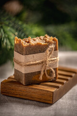 Piece of handmade natural soap on a wooden soap dish and a linen tablecloth against the background of green branches of the Christmas tree