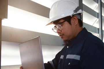 Chief engineer in hard hat walks through modern factory lighting while holding laptop. Successful handsome man in modern industrial environment.
