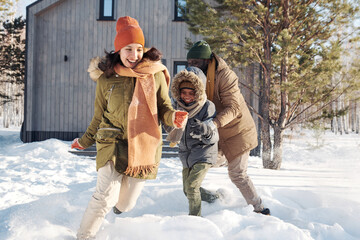 Young laughing woman in winterwear running along snowdrift while playing with her husband and son during winter vacation