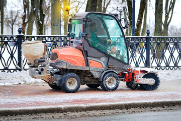 Tractor with rotating brush sweep snow from sidewalk. Small tractor clear snow from footpath, snow cleaning machine removing snow from pedestrian walkway