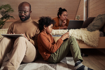 Cute boy with smartphone sitting on the floor next to his father using tablet while networking or...