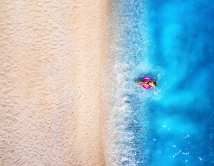 Aerial view of a woman swimming with pink swim ring in blue sea, empty sandy beach at sunset in...