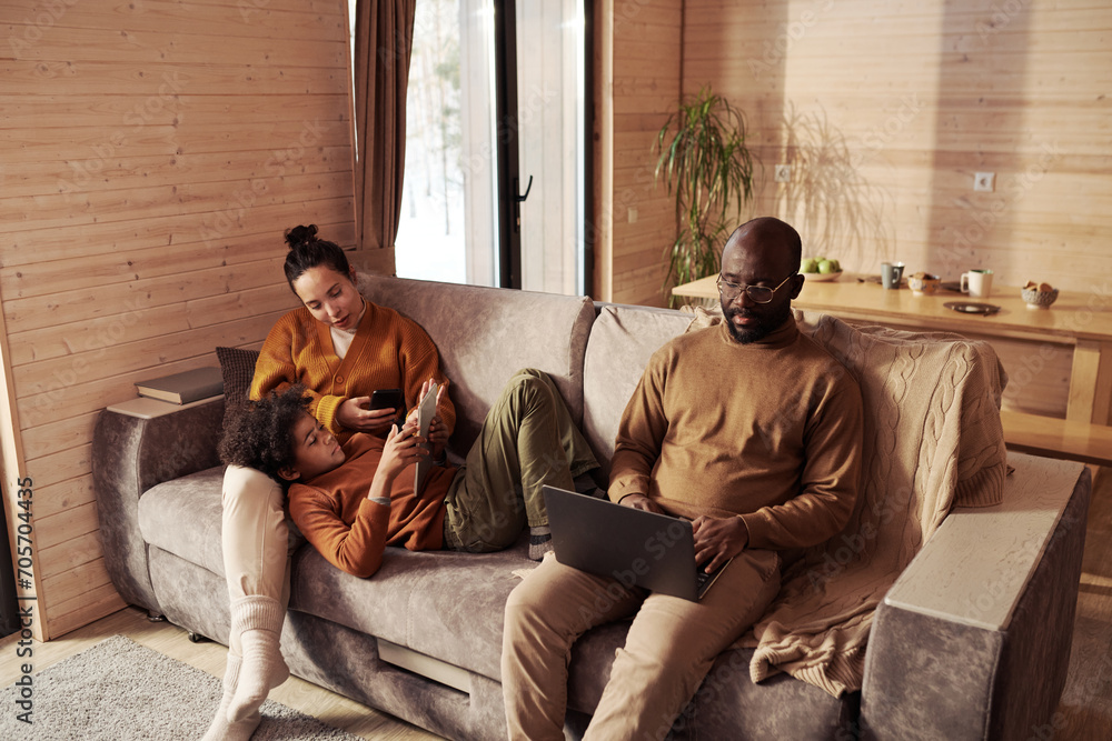 Wall mural Boy with tablet lying on couch next to his mother with mobile phone while serious young African American man using laptop during work