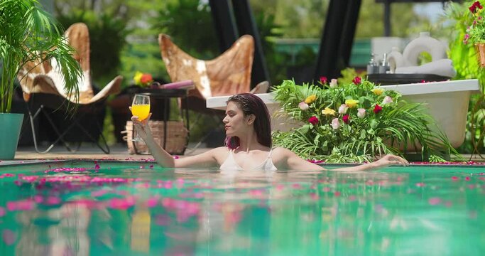 Indian Asian Hindu relax woman bathing rose petals water park pool edge resort hold glass pick up drink fresh juice happy young adult female girl luxury hotel villa trip do pose enjoy fun joy day