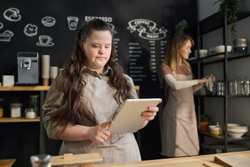 Young waitress with Down syndrome looking at tablet screen while standing by counter in front of...