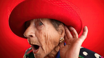 Funny portrait of smiling happy crazy elderly woman with no teeth, puts hand to ear to listen to a...