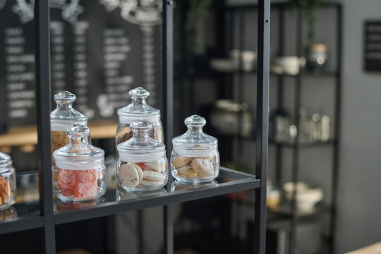 Group of jars containing macaroons and other sweets standing on shelf of large display in modern cafe against workplace of waiter or clerk