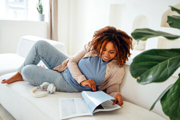 oung african woman sitting on sofa indoors at home while reading magazine or book
