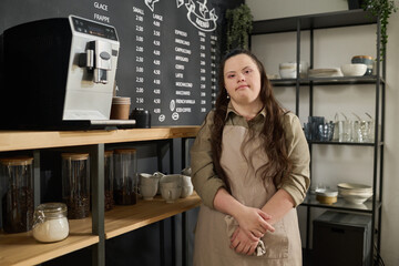 Young woman with Down syndrome wearing apron looking at camera while standing by coffee machine against blackboard with menu