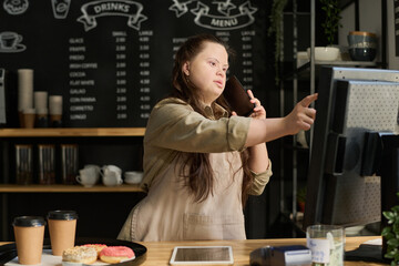Young waitress with mental disability talking on smartphone with client and pointing at computer...