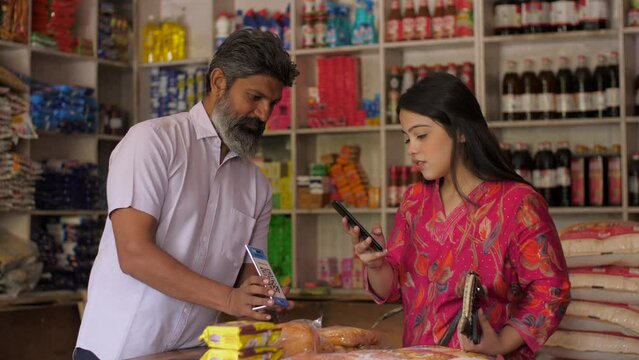 An Indian woman making an online payment at Kirana store - shopping concept  lifestyle  grocery store. A shopkeeper selling at his store - woman paying through a scanner or QR code  mode of payment...