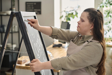 Young female worker of cafe with mental disability writing down menu with white chalk on blackboard while standing in front of camera