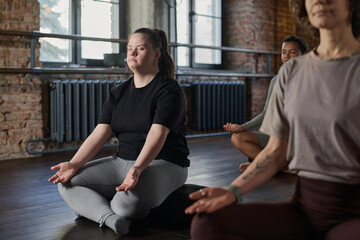 Young active woman with Down syndrome sitting in pose of lotus with her eyes closed and meditating...