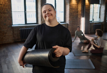 Young smiling sportswoman with Down syndrome holding rolled mat and looking at camera while...