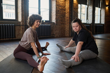 Young female fitness trainer and girl with Down syndrome sitting on mats in gym and doing physical exercise with outstretched legs