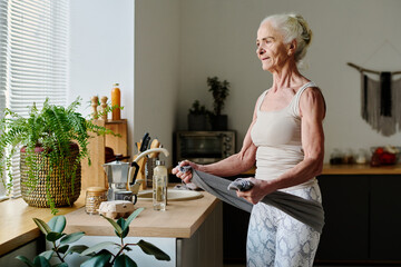 Healthy female boomer in activewear exercising with towel while standing by kitchen counter...