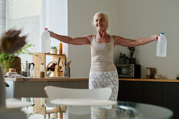 Smiling senior woman in activewear outstretching her arms and holding two plastic canisters with water while exercising in the kitchen - Powered by Adobe
